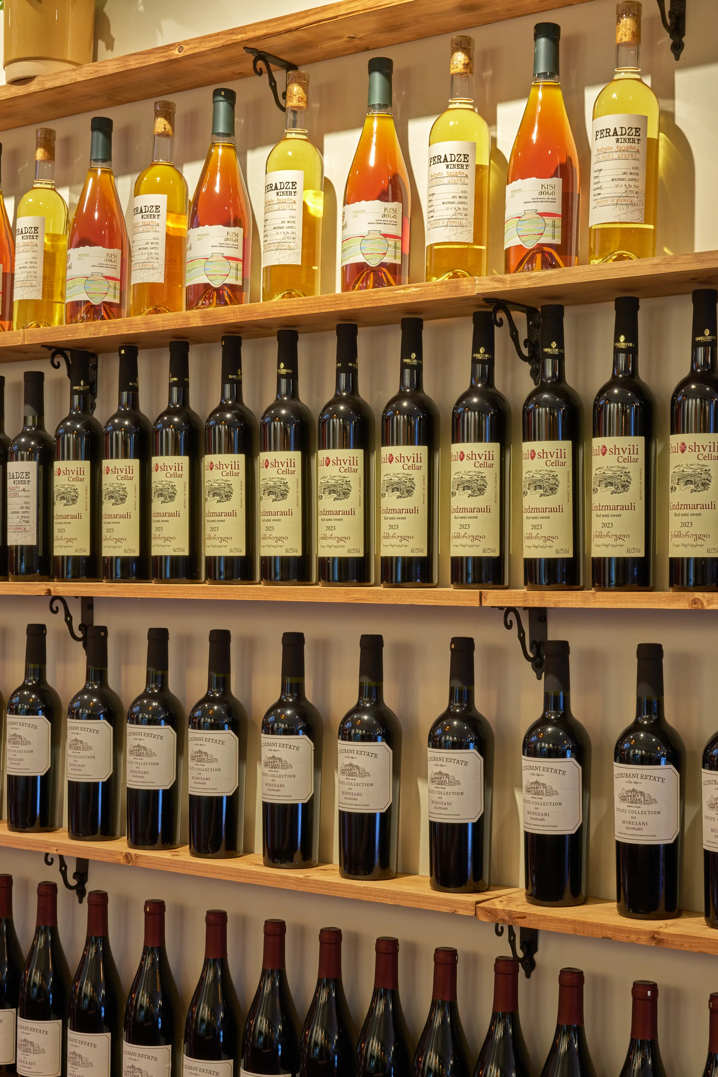 Close-up of amber and red wine bottles on wooden shelves, with labels from Peradze, Shaloshvili and Vazisubani